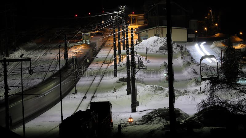 Night motion photo of a train passing a train station in winter. Electrified railway track. Illuminated station building and platform. Motion photography. Slow shutter speed. Night motion photo of a train passing a train station in winter. Electrified railway track. Illuminated station building and platform. Motion photography. Slow shutter speed.