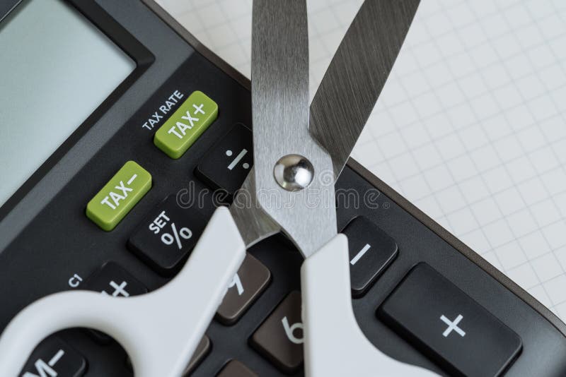 Tax cut, budget reduction, debt cutout concept, white scissors on black calculator with green button with tax minus and tax plus label on grid line paper note book. Tax cut, budget reduction, debt cutout concept, white scissors on black calculator with green button with tax minus and tax plus label on grid line paper note book