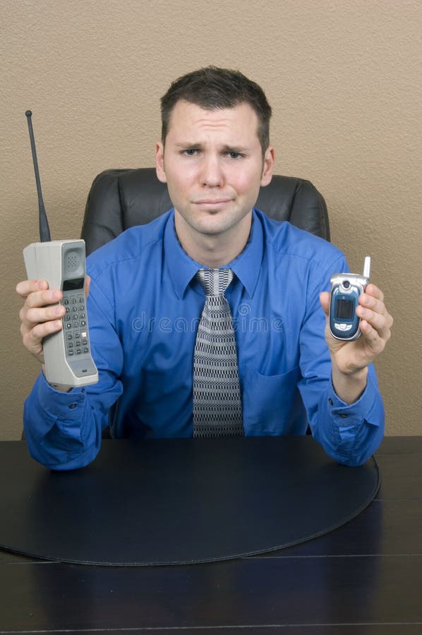 Guy in his office makes a funny face when comparing an old, brick-like early cell-phone to a minuscule nowadays flip phone. Guy in his office makes a funny face when comparing an old, brick-like early cell-phone to a minuscule nowadays flip phone