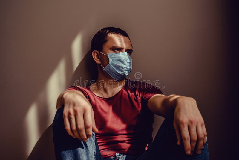 医用面具中令人伤感的成年男子肖像坐在墙上，上面有光影图案 在家保持隔离 免版税图库摄影