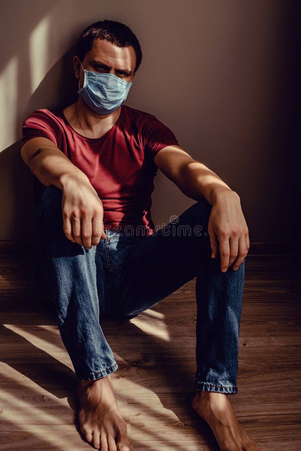 医用面具中心的可怜男人，坐在镶木地板上，上面有条纹形的光影图案 在家保持隔离 库存照片