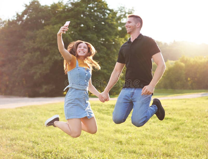 千禧一代的快乐情侣在夕阳休闲概念中一起跳起来，在一个年轻快乐的公园里自拍 免版税库存图片