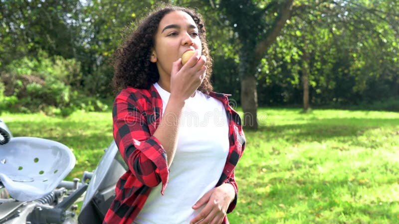 吃苹果的两种人种的非裔美国人的混合的族种十几岁的女孩年轻女人由一台拖拉机在果树园