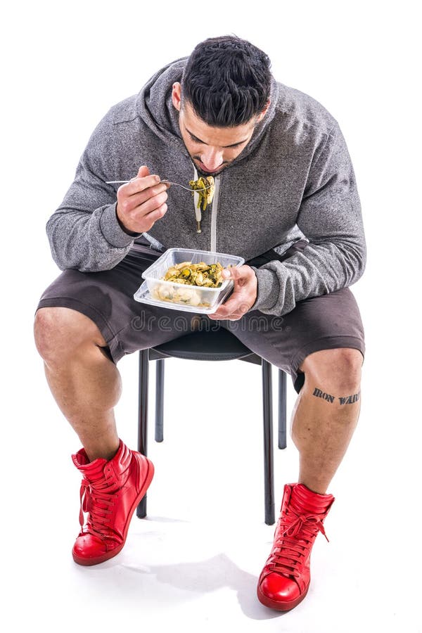 Hungry muscular young man gulping down food looking at plastic container without pausing as he takes another mouthful. Hungry muscular young man gulping down food looking at plastic container without pausing as he takes another mouthful
