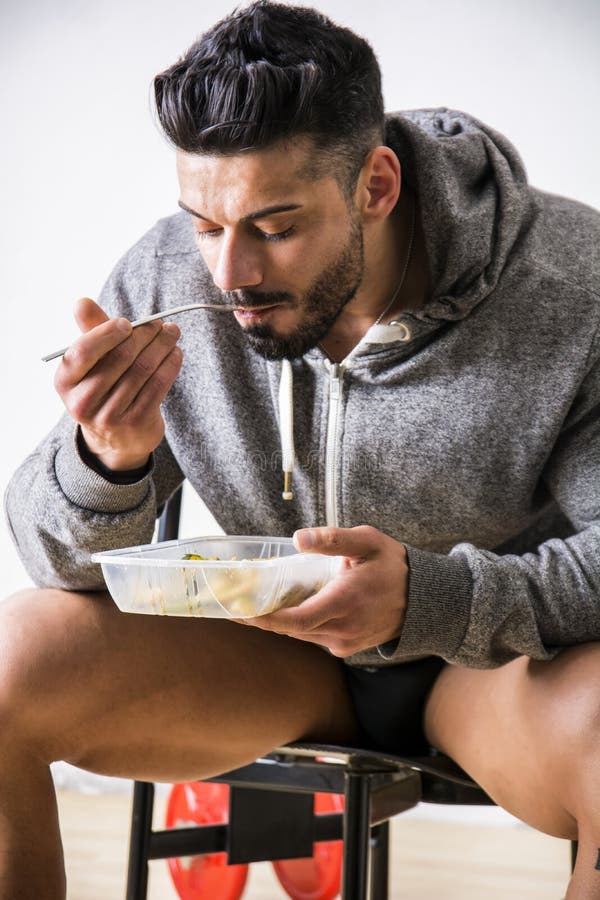 Hungry muscular young man gulping down food looking at plastic container without pausing as he takes another mouthful. Hungry muscular young man gulping down food looking at plastic container without pausing as he takes another mouthful