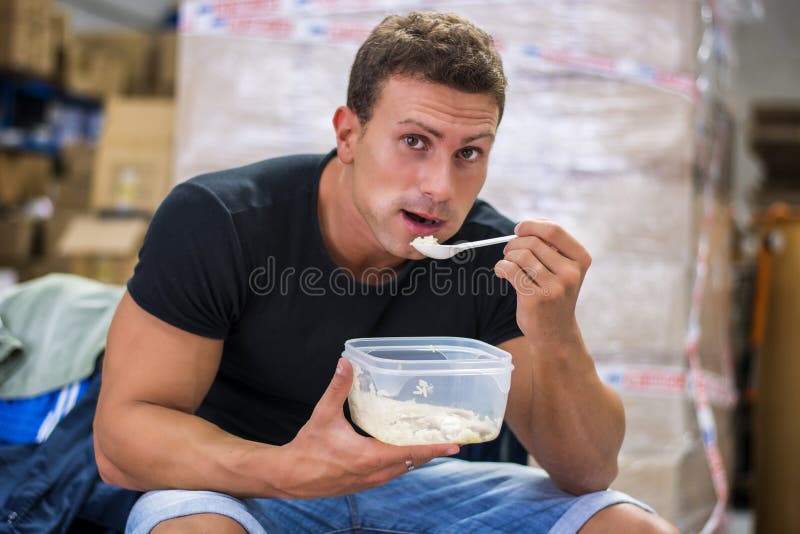 Hungry shirtless muscular young man gulping down food glancing across at the camera without pausing as he takes another mouthful. Hungry shirtless muscular young man gulping down food glancing across at the camera without pausing as he takes another mouthful