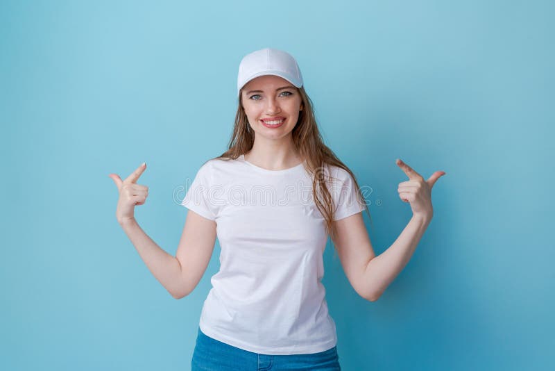 Hey look. Smiling beautiful female model pointing her fingers to an empty space and inviting to check it out showing an advertisement on a blue background. Dressed in white t-shirt and cap for mockup. Hey look. Smiling beautiful female model pointing her fingers to an empty space and inviting to check it out showing an advertisement on a blue background. Dressed in white t-shirt and cap for mockup