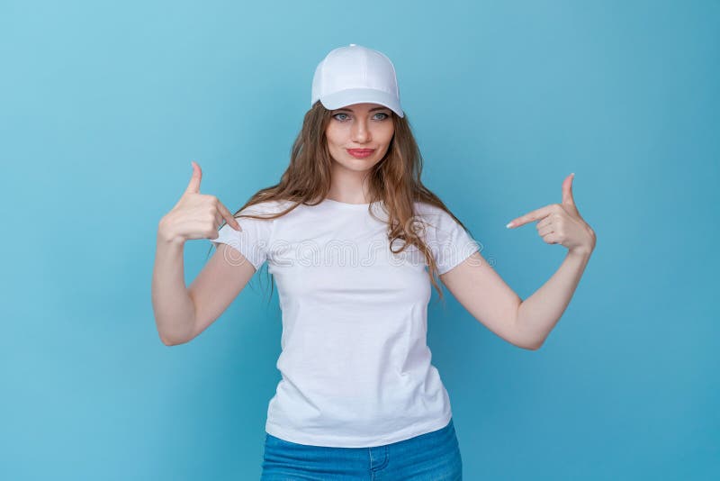 Hey look. Smiling beautiful female model pointing her fingers to an empty space and inviting to check it out showing an advertisement on a blue background. Dressed in white t-shirt and cap for mockup. Hey look. Smiling beautiful female model pointing her fingers to an empty space and inviting to check it out showing an advertisement on a blue background. Dressed in white t-shirt and cap for mockup