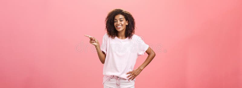 Hey look. Portrait of cute chill and friendly stylish african american woman with afro hairstyle holding hand on waist pointing right and smiling joyfully over pink background. Copy space. Hey look. Portrait of cute chill and friendly stylish african american woman with afro hairstyle holding hand on waist pointing right and smiling joyfully over pink background. Copy space