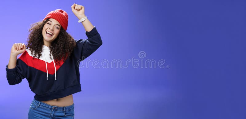 Hey come on dance with me. Friendly-looking bright and stylish cheerful woman with curly hairstyle wearing warm beanie raising hands up as enjoying great day, having fun outdoors over blue wall. Hey come on dance with me. Friendly-looking bright and stylish cheerful woman with curly hairstyle wearing warm beanie raising hands up as enjoying great day, having fun outdoors over blue wall.