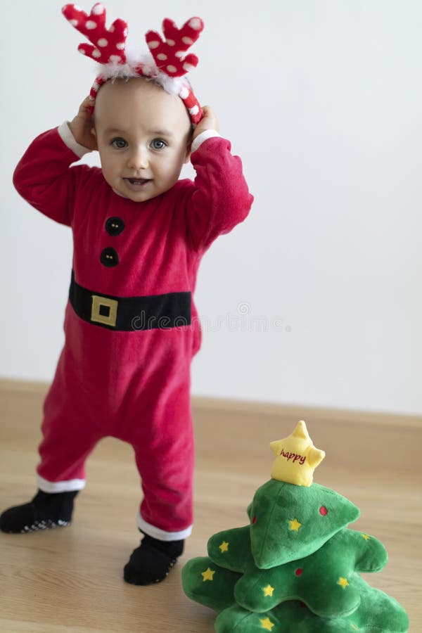 Baby girl wearing red Santa`s dress and deer horns behind small Christmas tree soft toy. First Christmas. Baby girl wearing red Santa`s dress and deer horns behind small Christmas tree soft toy. First Christmas