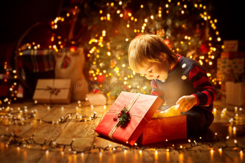 Christmas Child Open Present Gift, Happy Baby Boy looking to Magic Light in Box, Kid sitting front of Xmas Tree. Christmas Child Open Present Gift, Happy Baby Boy looking to Magic Light in Box, Kid sitting front of Xmas Tree