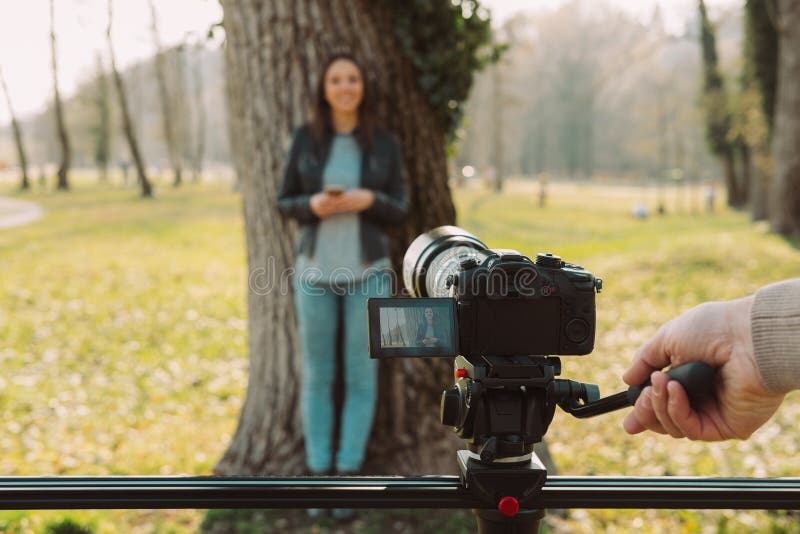 Video shooting at the park: video camera and operator hand on foreground and female model standing in the background. Video shooting at the park: video camera and operator hand on foreground and female model standing in the background