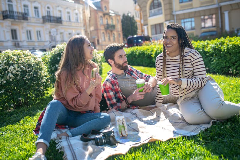Time relax. Three young satisfied friends at a picnic in a city square sitting on a plaid with coffee talking. Time relax. Three young satisfied friends at a picnic in a city square sitting on a plaid with coffee talking
