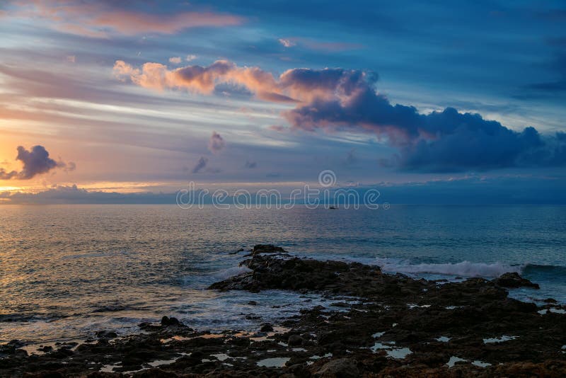 在宁静的海洋地平线上，夕阳在岩石海岸上，波涛缓和，还有远方的帆船 库存照片