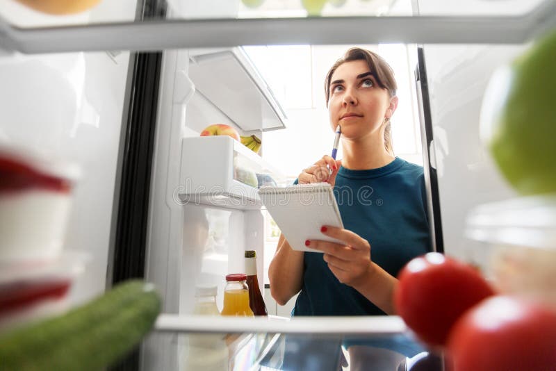 Healthy eating and diet concept - woman opening fridge and making list of necessary food at home kitchen. Healthy eating and diet concept - woman opening fridge and making list of necessary food at home kitchen