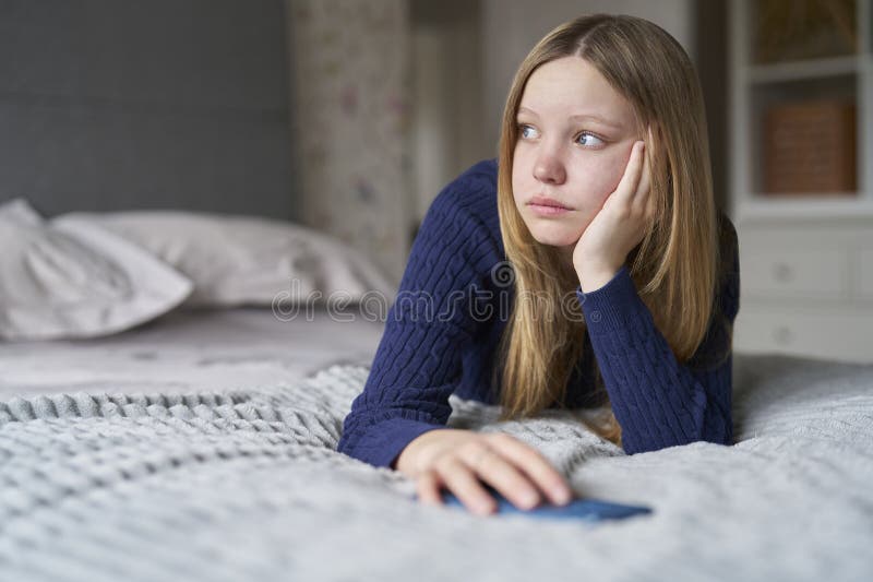 Unhappy Teenage Girl With Mobile Phone Lying On Bed At Home Anxious About Social Media Online Bullying And Using Phone Too Much. Unhappy Teenage Girl With Mobile Phone Lying On Bed At Home Anxious About Social Media Online Bullying And Using Phone Too Much