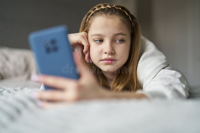 Unhappy Teenage Girl With Mobile Phone Lying On Bed At Home Anxious About Social Media Online Bullying And Using Phone Too Much. Unhappy Teenage Girl With Mobile Phone Lying On Bed At Home Anxious About Social Media Online Bullying And Using Phone Too Much