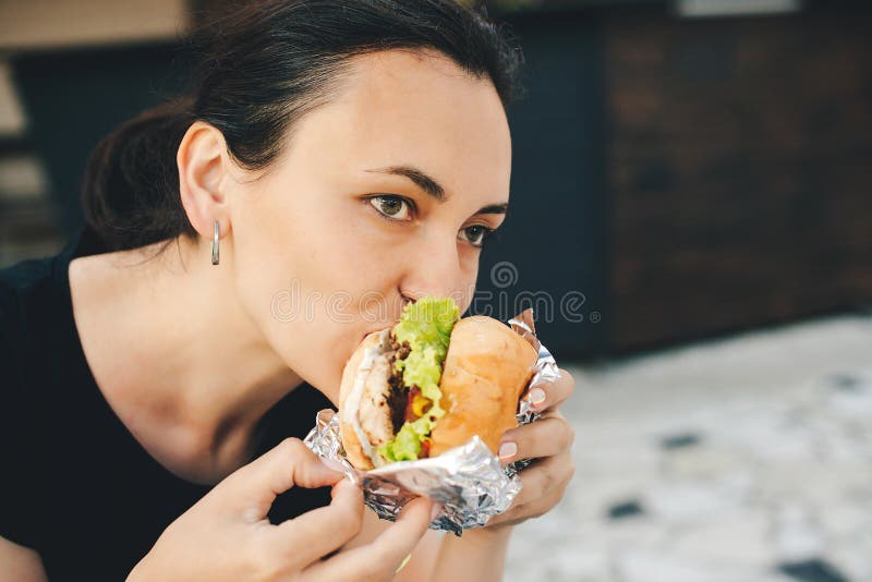 在户外咖啡馆吃美味汉堡的年轻女人 街头美食节 女孩喜欢快餐 不健康的饮食 库存图片 在户外咖啡馆吃美味汉堡的年轻女人 街头美食节 女孩喜欢快餐 不健康的饮食 库存图片
