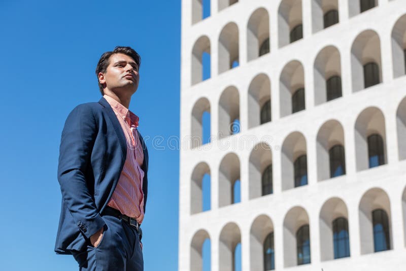 在户外穿衬衫穿夹克的商人. 手放在口袋里. 在他身后的一栋建筑和蓝天. 观察距离