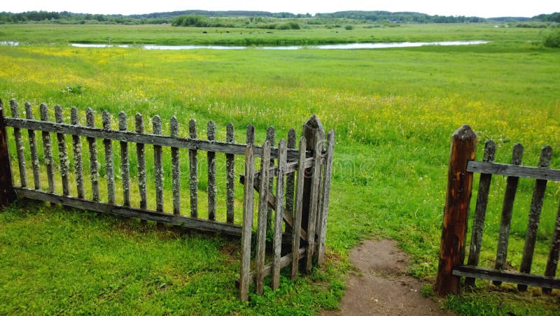 An open gate in a wooden fence and a green meadow beyond it, the path in the frame . Cloudy summer or late spring outside the city. An open gate in a wooden fence and a green meadow beyond it, the path in the frame . Cloudy summer or late spring outside the city