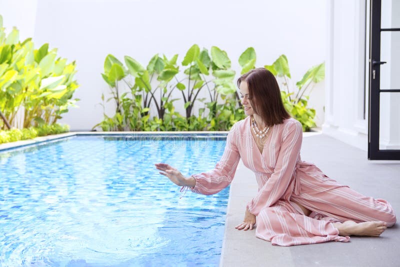 Beautiful brunette Caucasian woman wearing pajamas, while patting at the water surface playfully on the edge of swimming pool. Beautiful brunette Caucasian woman wearing pajamas, while patting at the water surface playfully on the edge of swimming pool