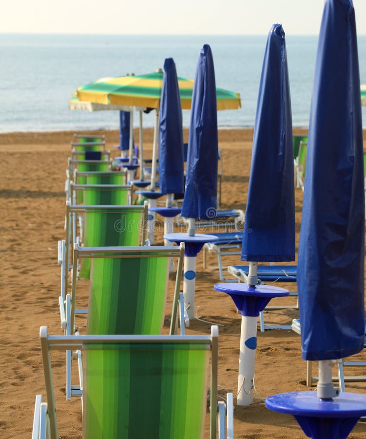 closed parasols with lounge chairs on the beach with no people in the early morning and the sea in the background. closed parasols with lounge chairs on the beach with no people in the early morning and the sea in the background