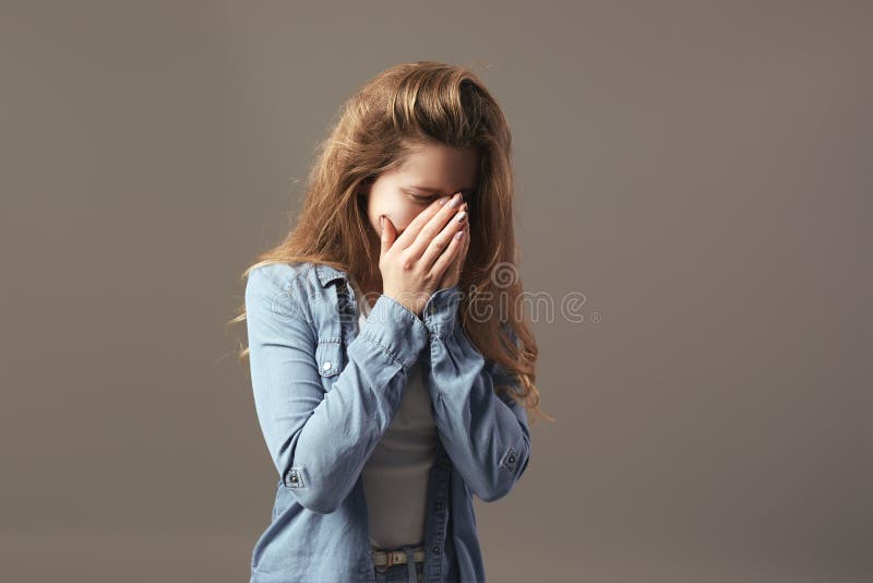 在白色T恤和斜纹布打扮的哀伤的棕色毛发的女孩握她的在她的面孔的手在灰色背景 免版税库存照片 在白色T恤和斜纹布打扮的哀伤的棕色毛发的女孩握她的在她的面孔的手在灰色背景 免版税库存照片
