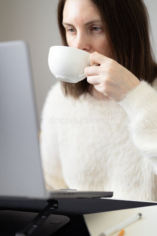 Focused woman drinking coffee in front of laptop screen. Using laptop stand for comfortable working. Busy businesswoman, home office, online meeting, shopping online, overtime working. Focused woman drinking coffee in front of laptop screen. Using laptop stand for comfortable working. Busy businesswoman, home office, online meeting, shopping online, overtime working