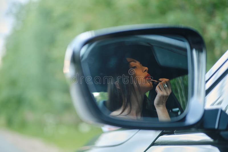 坐在汽车里涂唇膏的女子的后视镜中的反射 免版税库存照片