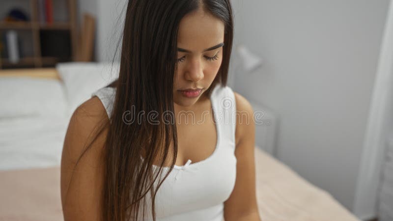 坐在舒适卧室卧室的女人，有一个年轻的、漂亮的西语裔黑发姑娘，长发穿着休闲装 库存图片