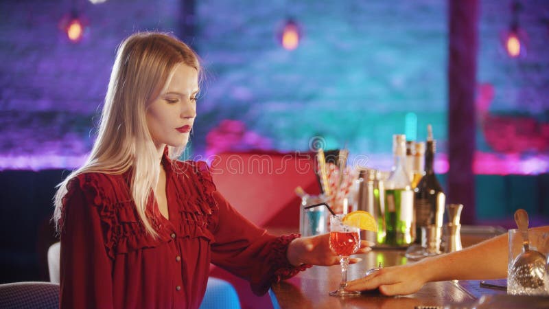 Gorgeous young woman sitting by the bartender stand - taking the drink from the stand and takes away the straw. Mid shot. Gorgeous young woman sitting by the bartender stand - taking the drink from the stand and takes away the straw. Mid shot