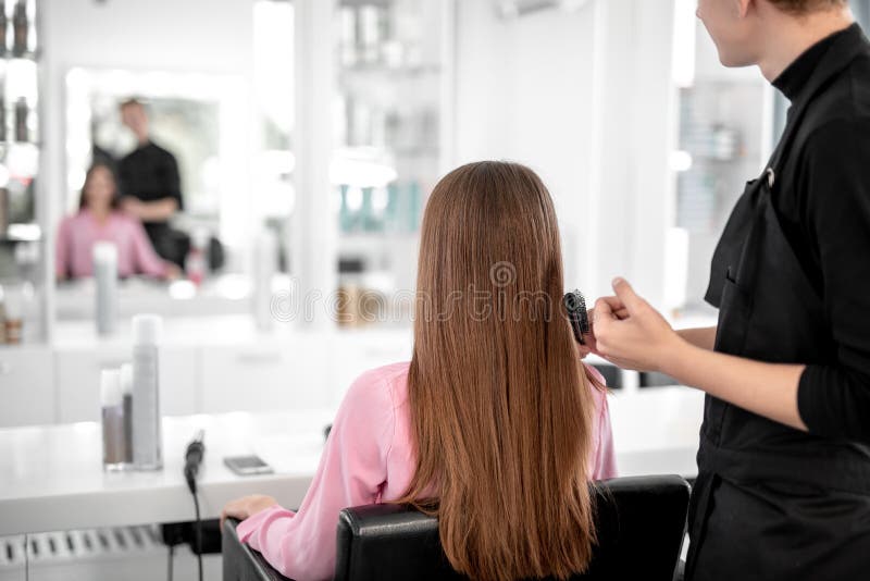 Long haired women sitting and looking at her reflection while professional hairdresser standing near and brushing her hair. Long haired women sitting and looking at her reflection while professional hairdresser standing near and brushing her hair
