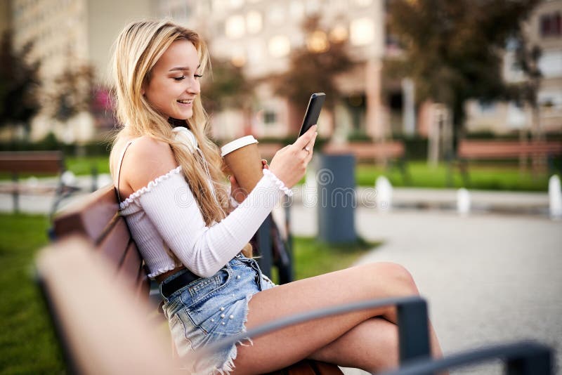 坐在长椅上、在智能手机上闲聊、一边拿着咖啡，一边拿着纸杯 免版税库存图片