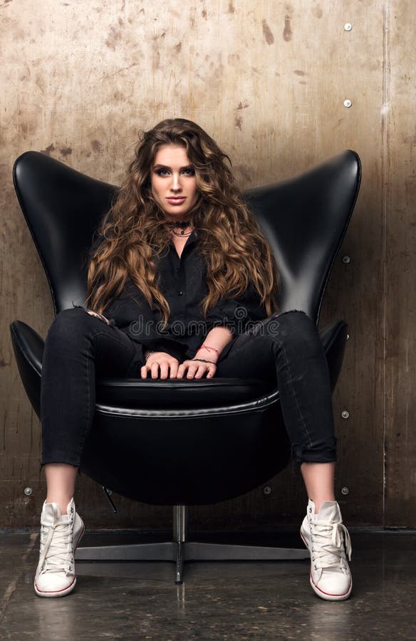 Vertical portrait of a domineering young woman sitting in the black chair. Black clothes and white sneakers. Luxurious long wavy hair. Vertical portrait of a domineering young woman sitting in the black chair. Black clothes and white sneakers. Luxurious long wavy hair