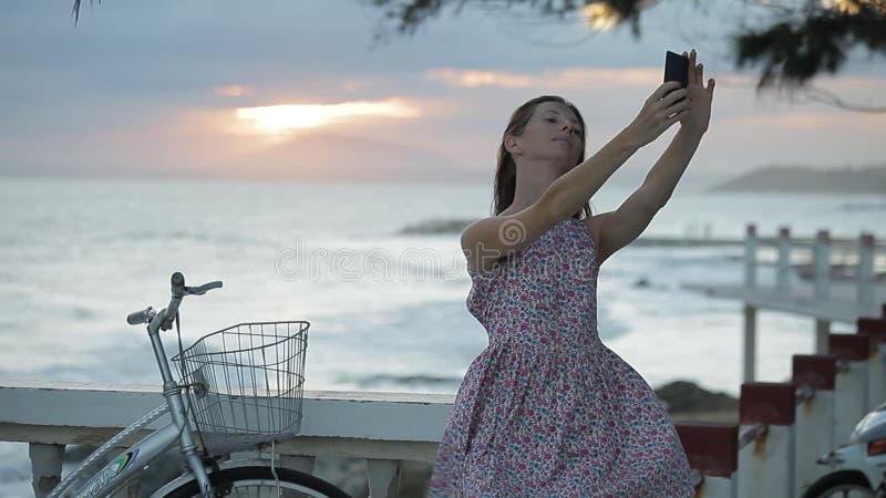 夏天礼服的女孩在堤防做selfie靠近海滩在日落