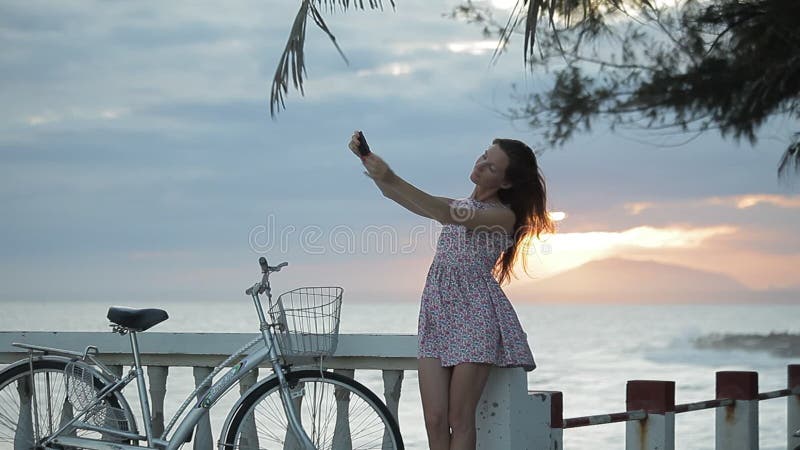 夏天礼服的女孩在堤防做selfie靠近海滩在日落