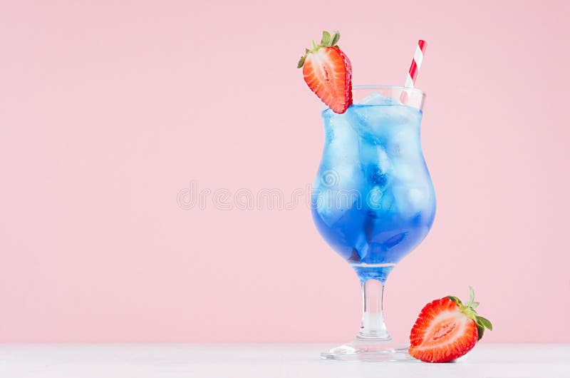 Summer blue alcohol drink with curacao liqueur, ice cubes, strawberry slice, straw in glamour glass on soft light pink background. Summer blue alcohol drink with curacao liqueur, ice cubes, strawberry slice, straw in glamour glass on soft light pink background