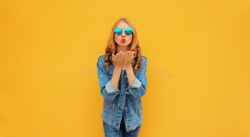 夏日时尚的年轻美女吹嘴唇向天空发出甜美的吻，戴着圆形太阳镜和牛仔夹克 库存图片