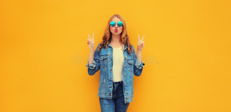 夏日时尚的年轻美女吹嘴唇向天空发出甜美的吻，戴着圆形太阳镜和牛仔夹克 库存照片