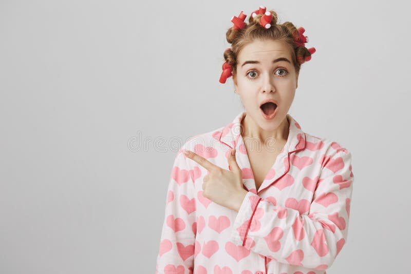 头发卷发的人和睡衣的惊奇的和震惊娘儿们女孩与心脏打印表明左与食指和 免版税库存照片 头发卷发的人和睡衣的惊奇的和震惊娘儿们女孩与心脏打印表明左与食指和 免版税库存照片