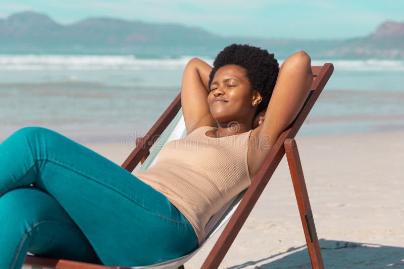 African american mature woman with hands behind head and eyes closed relaxing on deckchair at beach. summer, nature, afro hair, relaxation, unaltered, lifestyle, enjoyment and holiday concept. African american mature woman with hands behind head and eyes closed relaxing on deckchair at beach. summer, nature, afro hair, relaxation, unaltered, lifestyle, enjoyment and holiday concept.