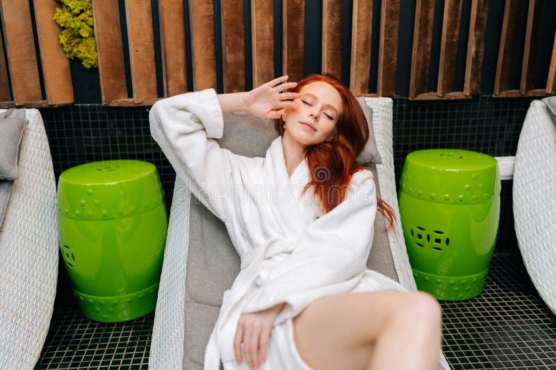 Portrait of pretty redhead young woman wearing white bathrobe stroking face lying with closed eyes on deckchair after spa treatment at beauty salon. Spa, sauna, skin care, lifestyle, wellness concept. Portrait of pretty redhead young woman wearing white bathrobe stroking face lying with closed eyes on deckchair after spa treatment at beauty salon. Spa, sauna, skin care, lifestyle, wellness concept.