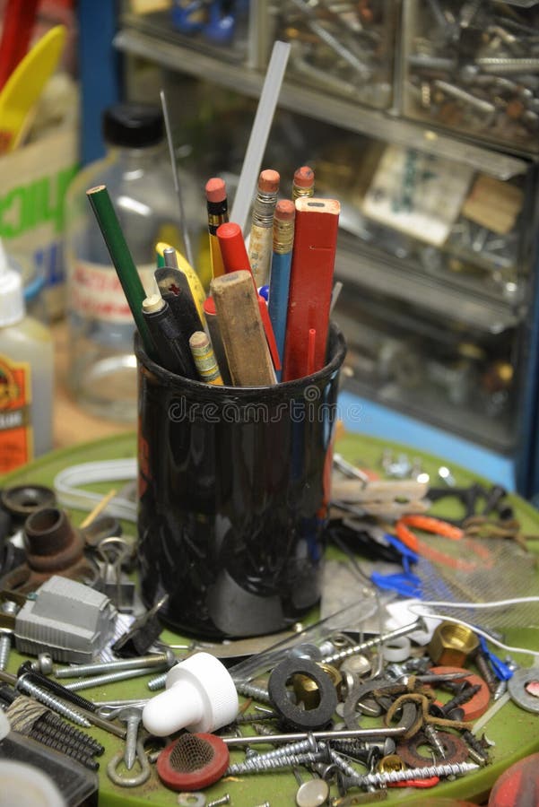 Pencil cup on the middle of a worktable full of various and assorted supplies.  Screws, washers, staples, new and old assorted bits and pieces to be helpful. Pencil holder.  Black mug. . Pencil cup on the middle of a worktable full of various and assorted supplies.  Screws, washers, staples, new and old assorted bits and pieces to be helpful. Pencil holder.  Black mug.