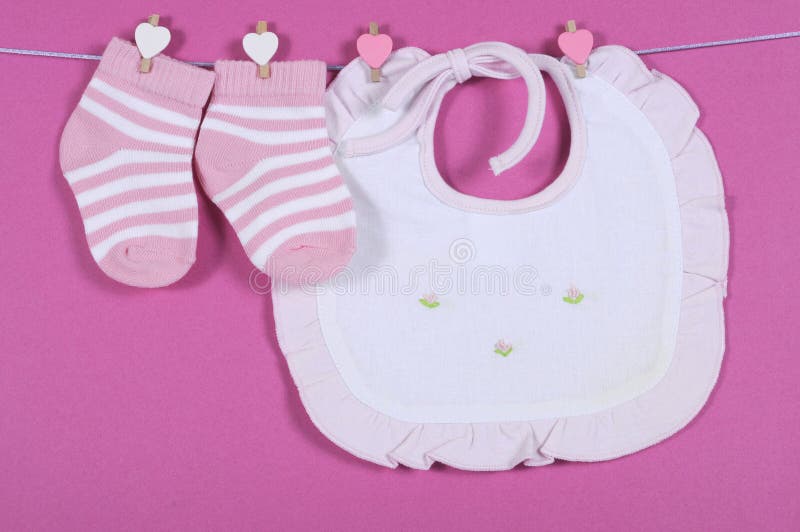 Baby girl nursery cute pink and white stripe socks and bib hanging from pegs on a line against a pink background for baby shower or newborn girl greeting card. Baby girl nursery cute pink and white stripe socks and bib hanging from pegs on a line against a pink background for baby shower or newborn girl greeting card.