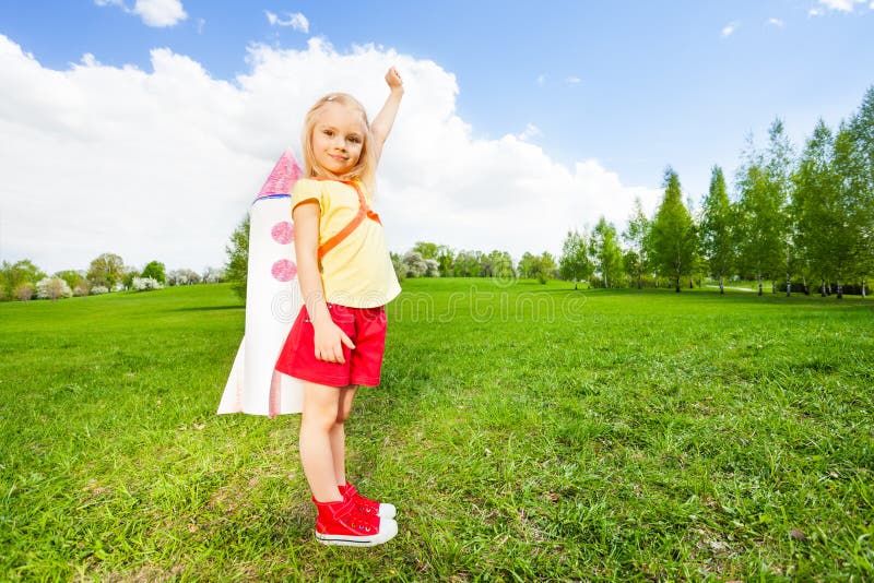 Girl wears paper rocket toy on her shoulders and holds arm straight up during sunny day in the green field. Girl wears paper rocket toy on her shoulders and holds arm straight up during sunny day in the green field