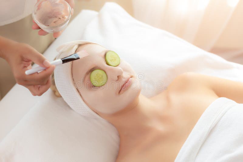 女按摩师或按摩治疗师应用的奶油色面具用途刷子于健康皮肤面孔的美丽的顾客面孔用黄瓜 免版税图库摄影 女按摩师或按摩治疗师应用的奶油色面具用途刷子于健康皮肤面孔的美丽的顾客面孔用黄瓜 免版税图库摄影