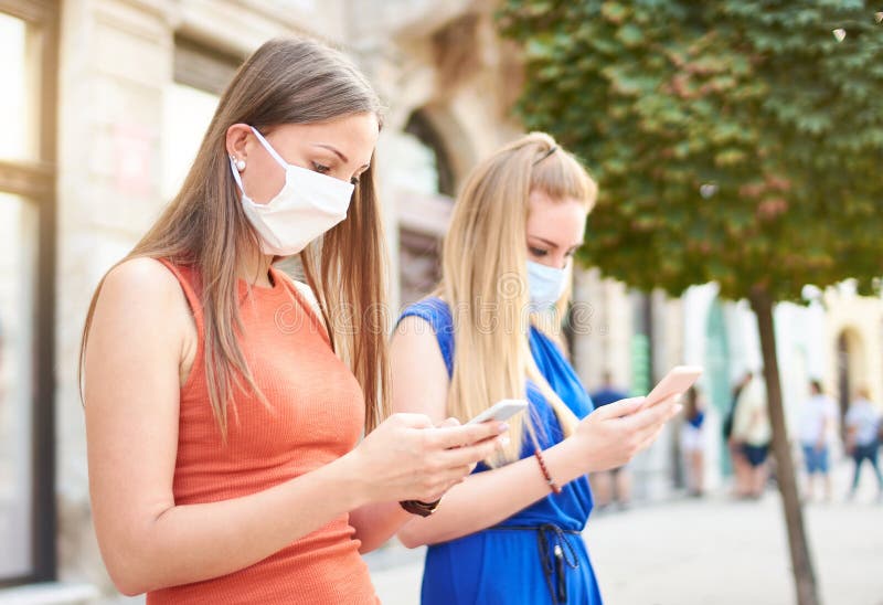 女朋友戴口罩使用智能手机年轻千禧一代查看社交媒体网络的更新新常态 免版税库存照片