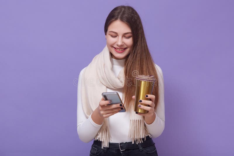 室内照片中,长发活泼的年轻女性手持智能手机和热杯,查看社交网络, 库存照片 室内照片中,长发活泼的年轻女性手持智能手机和热杯,查看社交网络, 库存照片