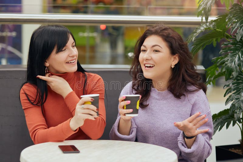 Indoor picture of two charismatic beautiful sweet brunettes sitting at table, holding paper cups with hot drink, talking actively, telling stories to each other, making gesture, having lunchtime. Indoor picture of two charismatic beautiful sweet brunettes sitting at table, holding paper cups with hot drink, talking actively, telling stories to each other, making gesture, having lunchtime